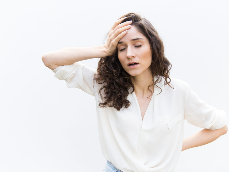 Exhausted tired woman with closed eyes touching head. Wavy haired young woman in casual shirt standing isolated over white background. Headache or burnout concept